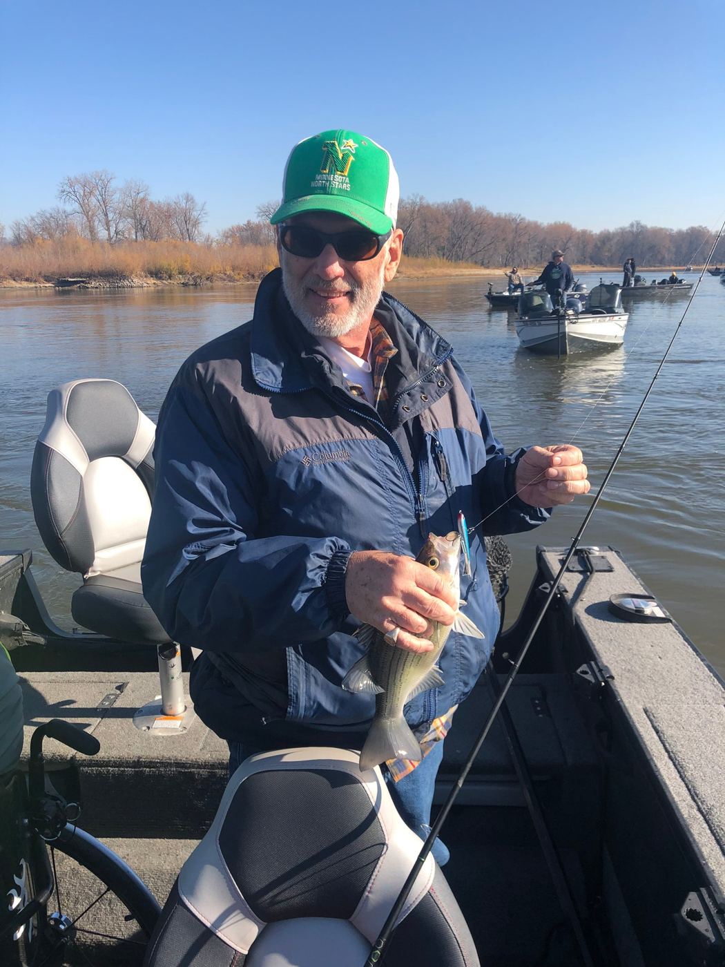 A smiling angler in a green cap holds up a fish while seated in a boat on the lake.