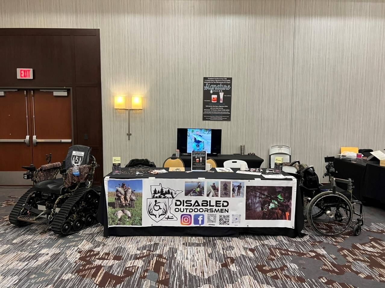 The Disabled Outdoorsmen MN outreach booth at an expo, with a banner, photos, and a tracked wheelchair on display.