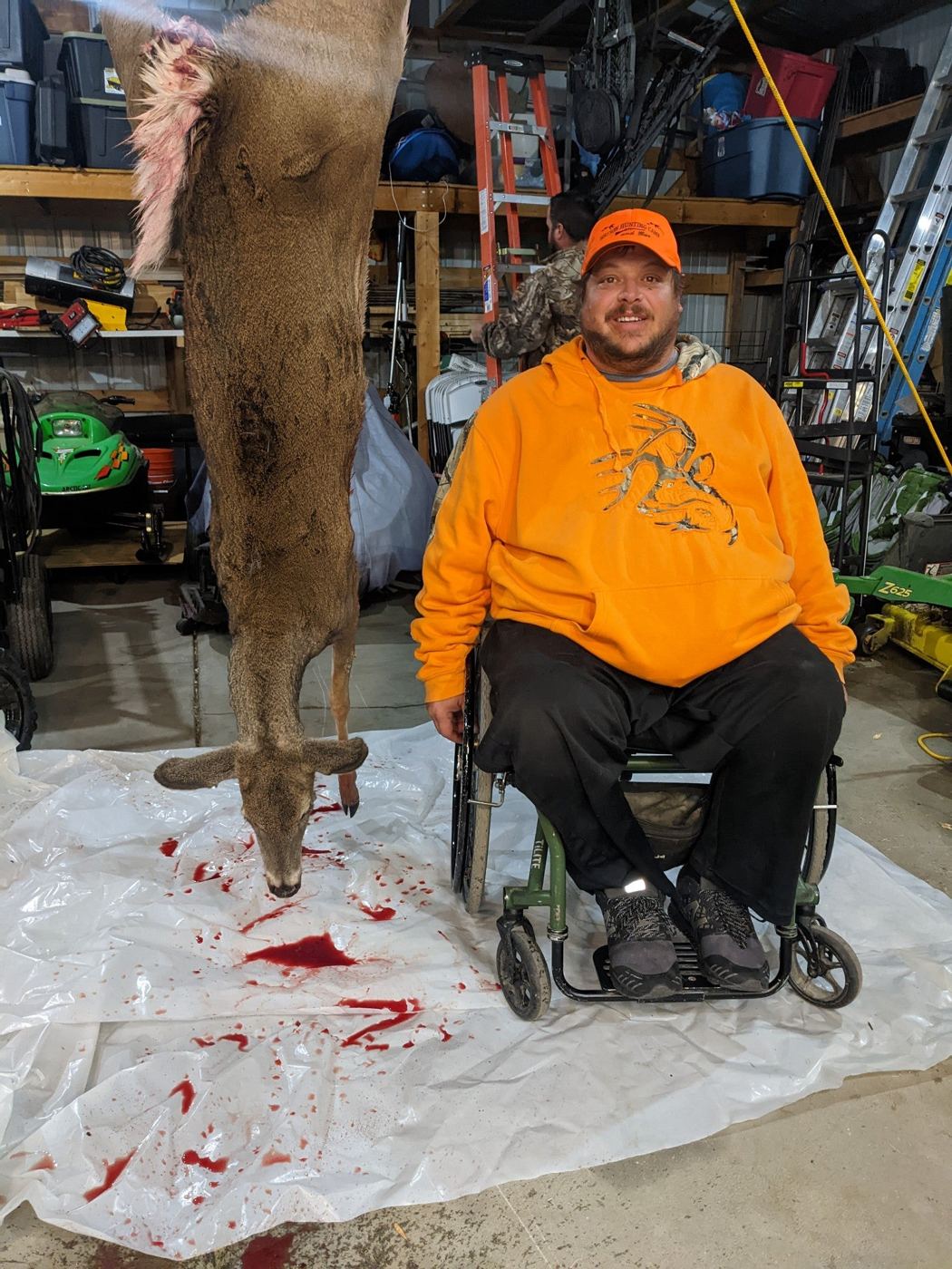 A hunter in an orange hoodie sits in a wheelchair in front of a hanging deer.