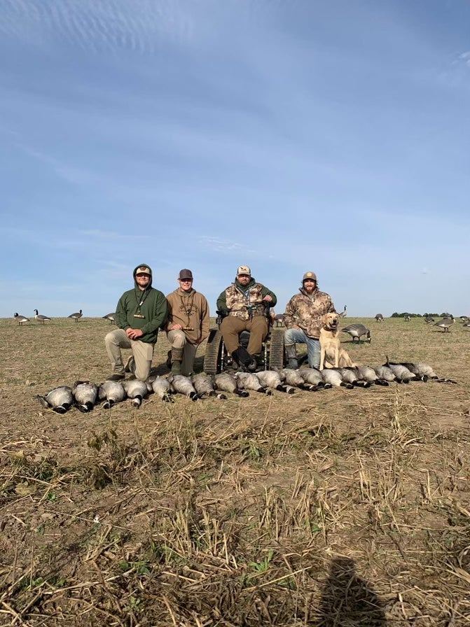 A group of hunters, including a participant in a wheelchair, stands behind a long line of geese in a harvested field.