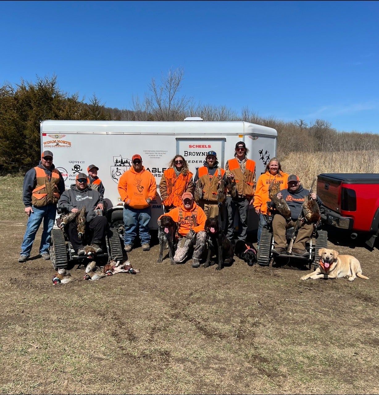 A large group of hunters and volunteers in blaze orange pose in front of the trailer with harvested birds.