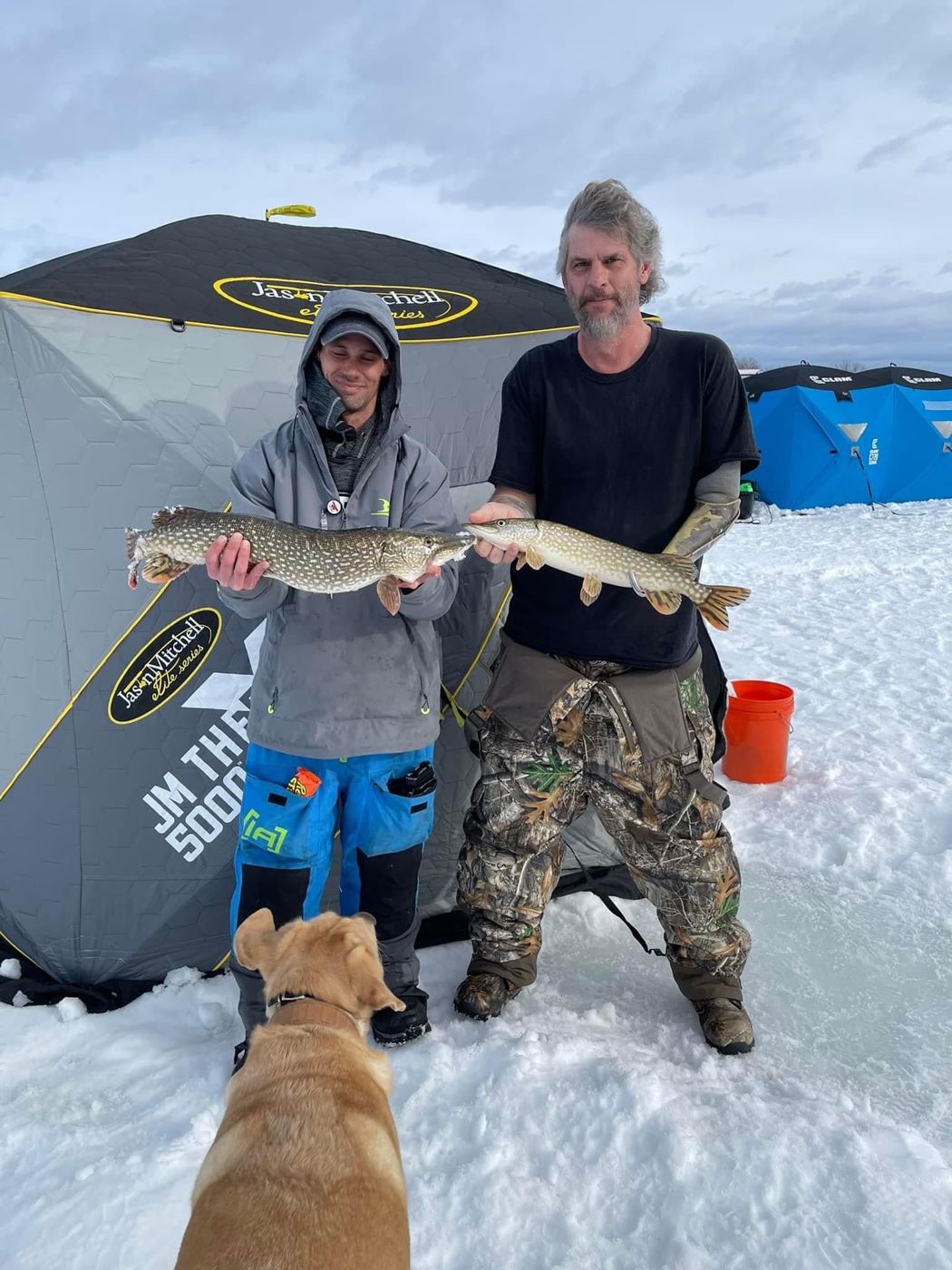 Two anglers on the ice show off their catch beside a portable fish house.