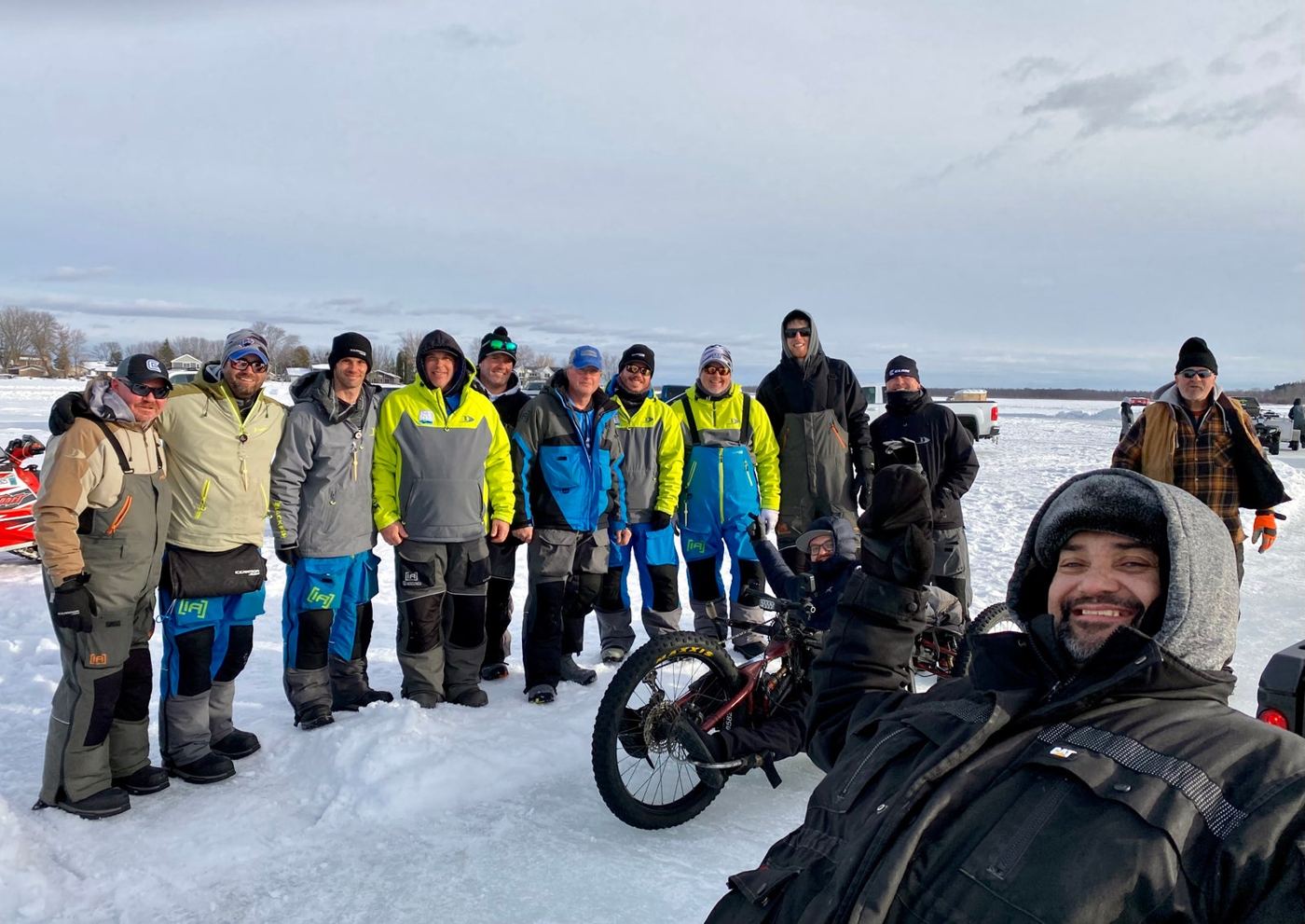 A large group of members and volunteers lined up on a frozen lake, with a participant in a tracked chair in front.
