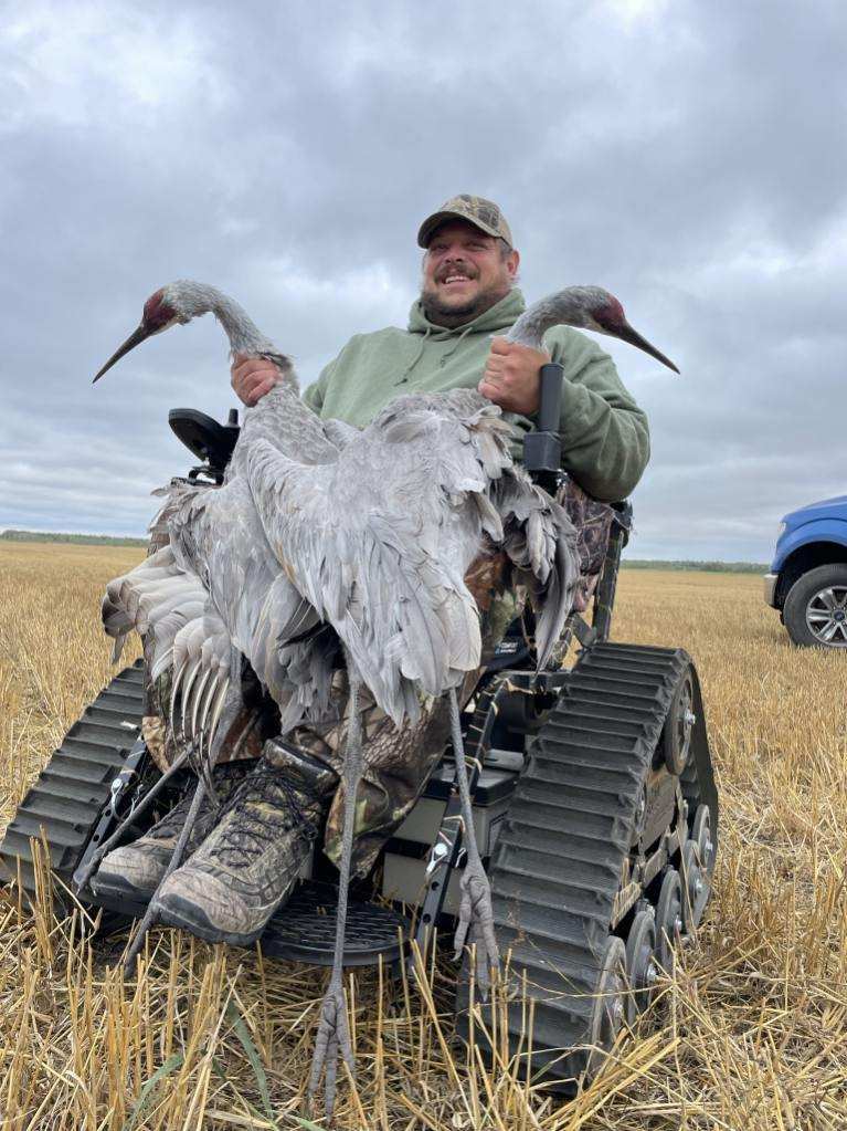 A hunter in a tracked all-terrain wheelchair holds up two sandhill cranes in a harvested field.