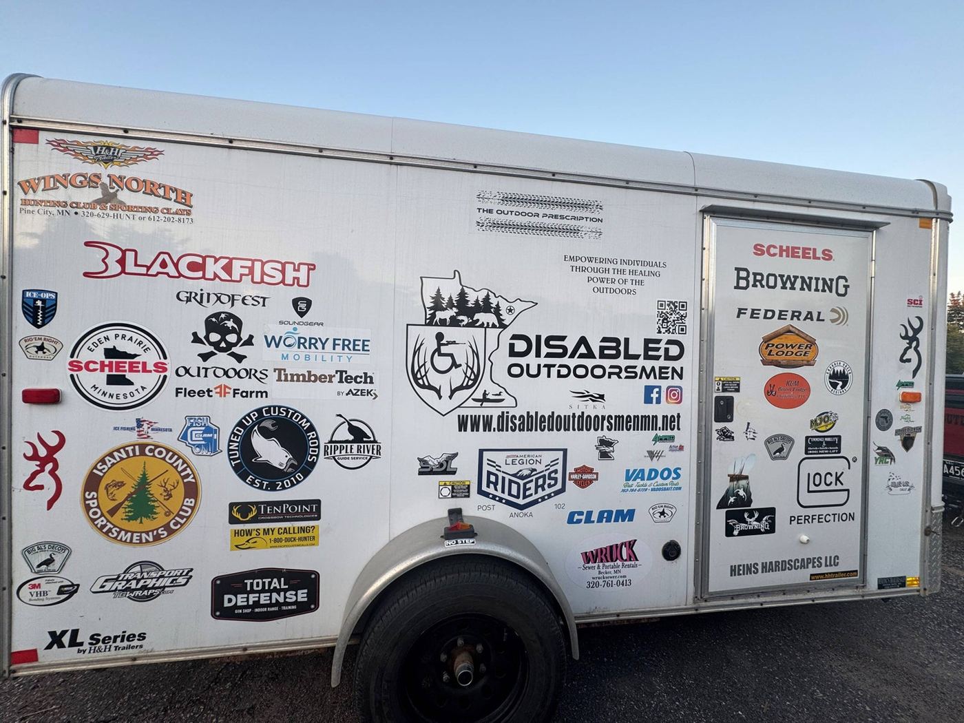 The Disabled Outdoorsmen MN equipment trailer, covered in sponsor logos, parked in a field.