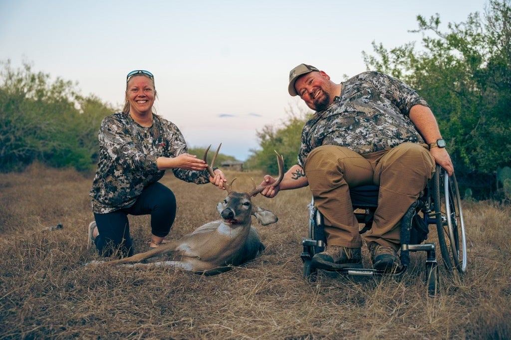 Two hunters, one in a wheelchair, smile together in a field at sunset.