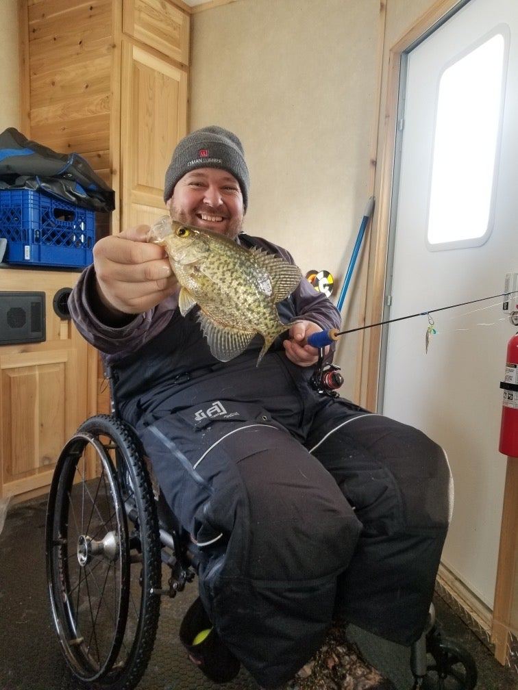 A man in a wheelchair inside a fish house proudly holds up a fish.