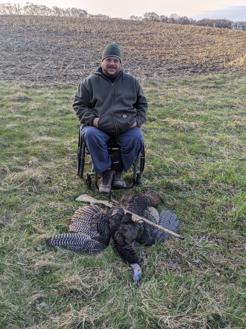 A hunter seated in a wheelchair in a grassy field with a harvested bird in front of him.